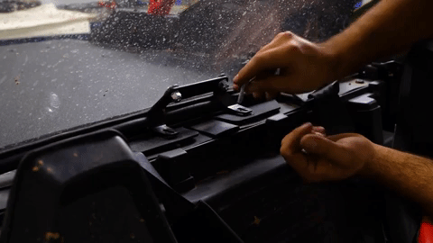 One hand holds a bolt in the palm while the other twists a similar bolt into place on the bracket along the rear windshield.