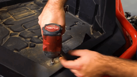 One hand holds a drill and the other hand collects the bolts as they are removed from the machine's bed.