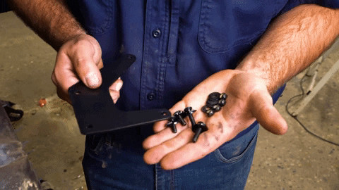 Braden holds a fistful of nuts, bolts, and washers on one hand and a U-shaped metal bracket in the opposite hand.