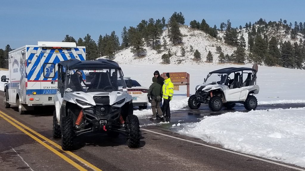 Two side-by-sides and an ambulance wait a trailhead along with a state trooper. There's a thick layer of snow on the ground.