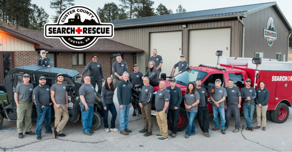 Multiple people stand in front of a building. They're all dressed in grey long sleeve shirts and there's a side-by-side and a rescue vehicle in the frame.