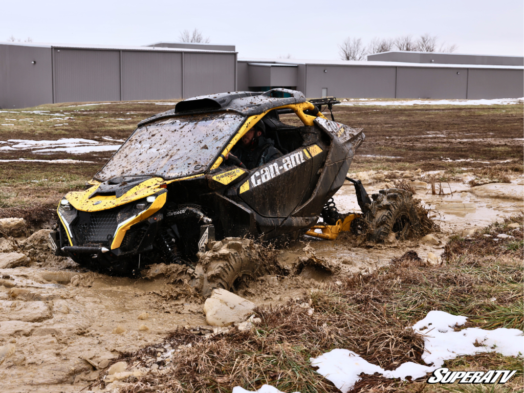 A Can-Am Maverick R plowing through a deep mud pit with debris all over the windshield.