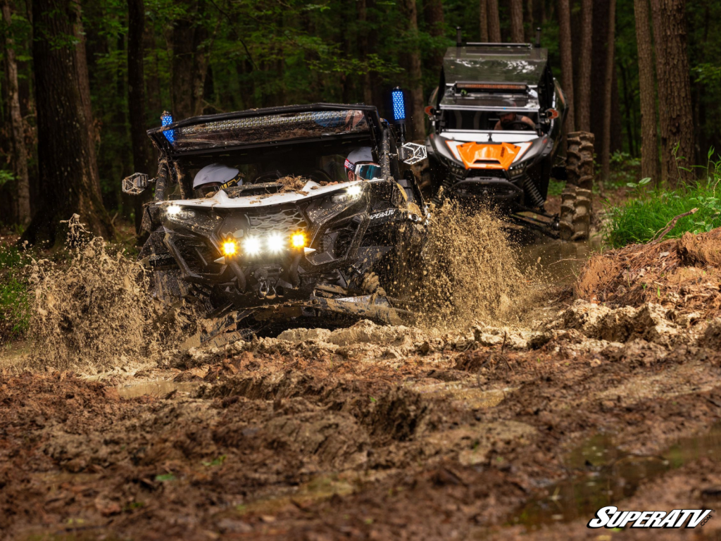 A pair of UTVs diving into a deep mud hole on a tree-lined trail.