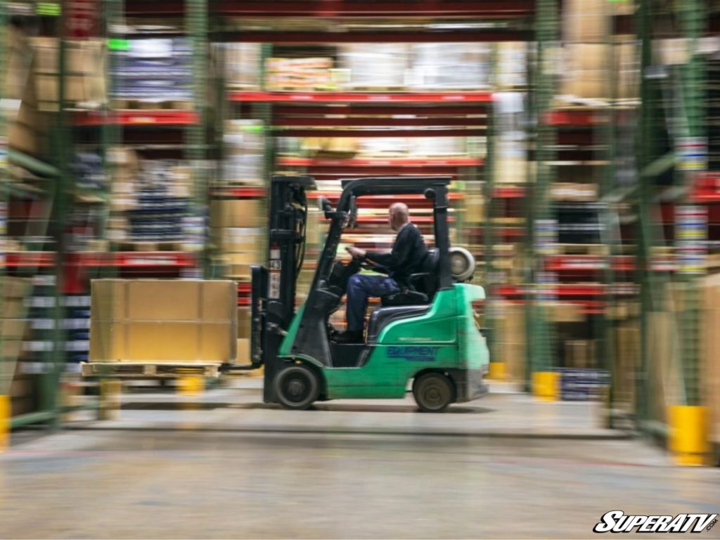 A man drives a green forklift through a SuperATV warehouse.
