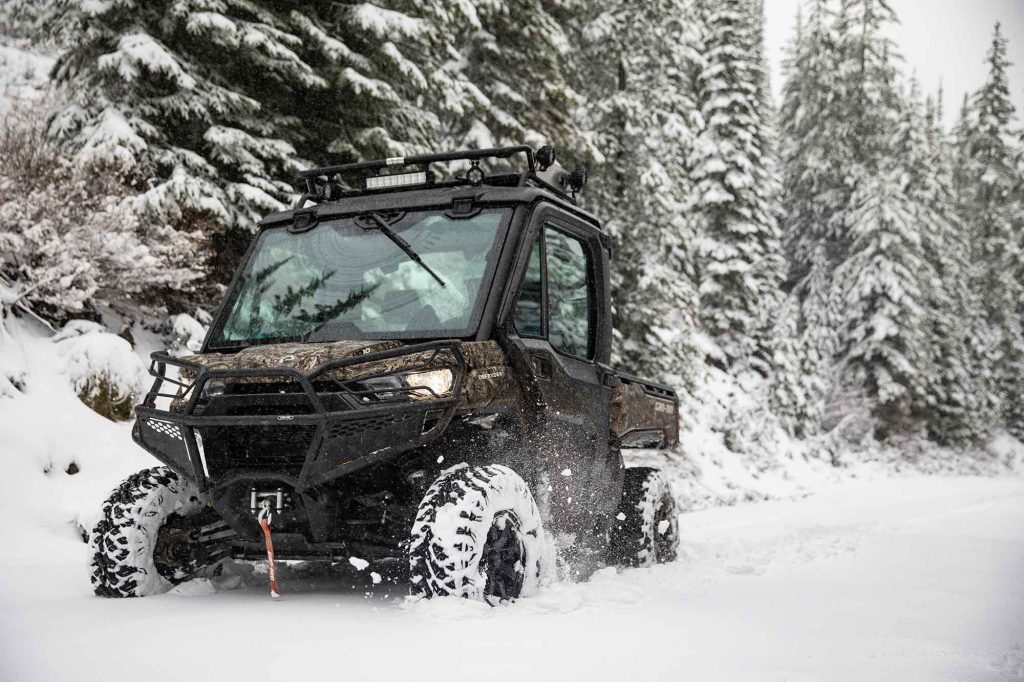 A Can-Am Defender in camo riding through deep snow on a forest trail.