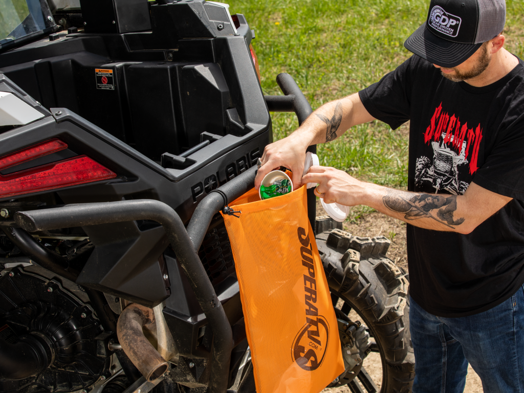 A man slides a handful of trash into an orange SuperATV-branded mesh trash bag.