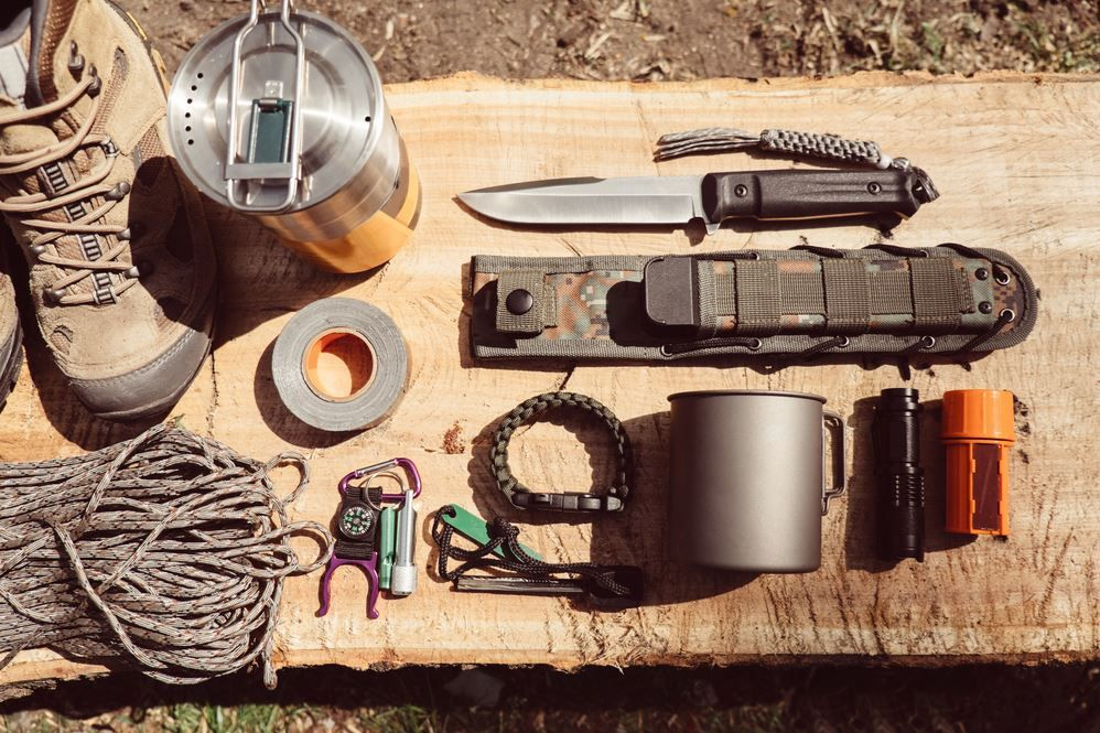 A series of off-road supplies laid out on a black of wood. There's an insulated container, a field knife, boots, and more.
