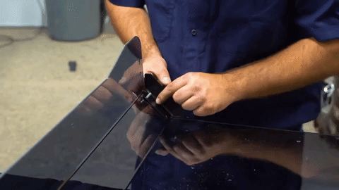 A man in a blue shirt tightens nuts on the back of a polycarbonate windshield with his thumb and forefinger. 