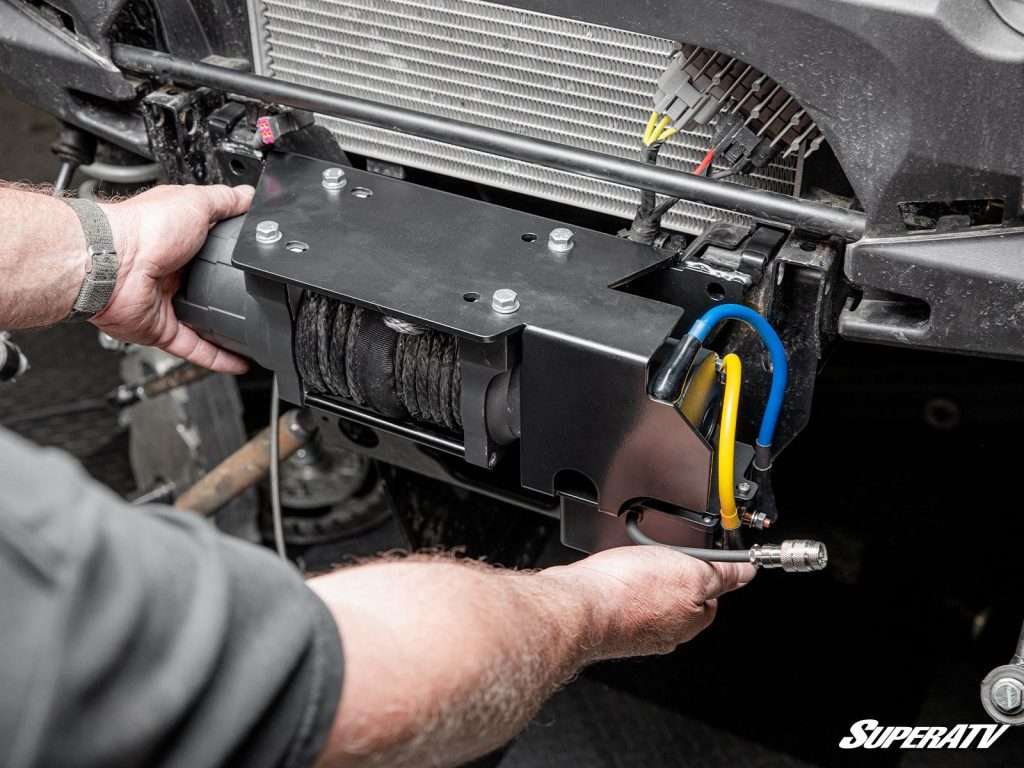 A man lifts a pre-wired winch into place in front of a machine's radiator.